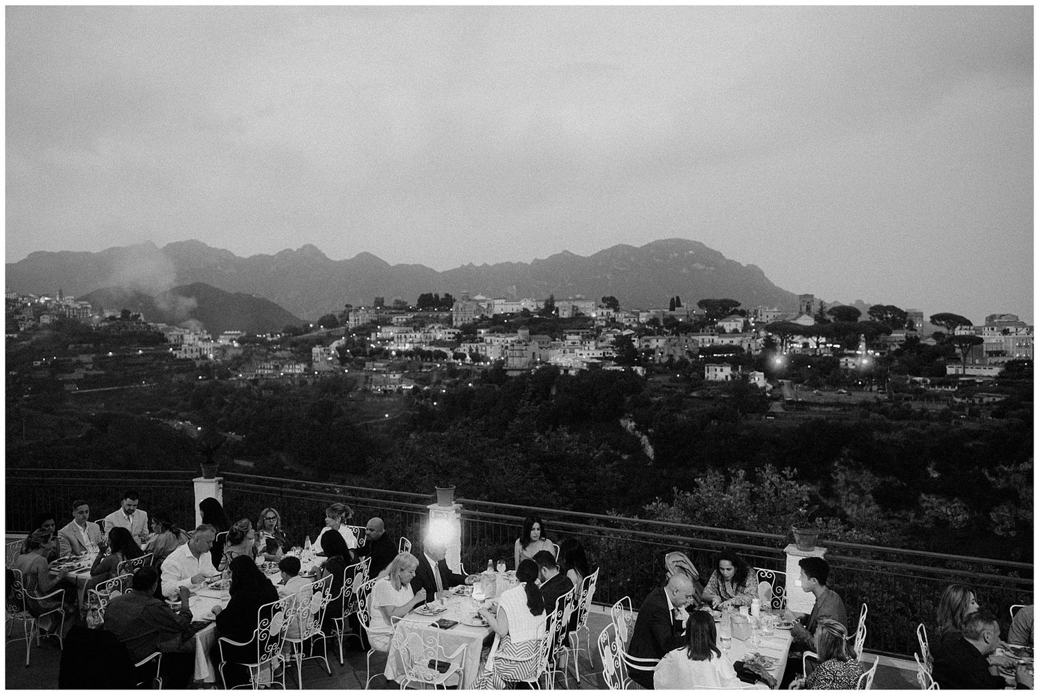 People gathered outdoors at a scenic viewpoint overlooking a mountainous town with houses dotting the hillside in black and white.