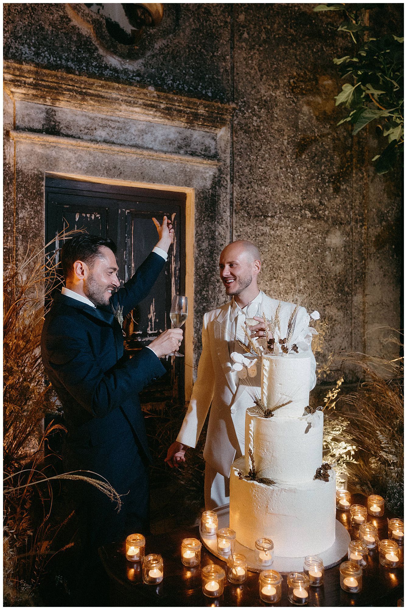 Two grooms share a celebratory moment by their elegant white wedding cake surrounded by romantic candlelight.