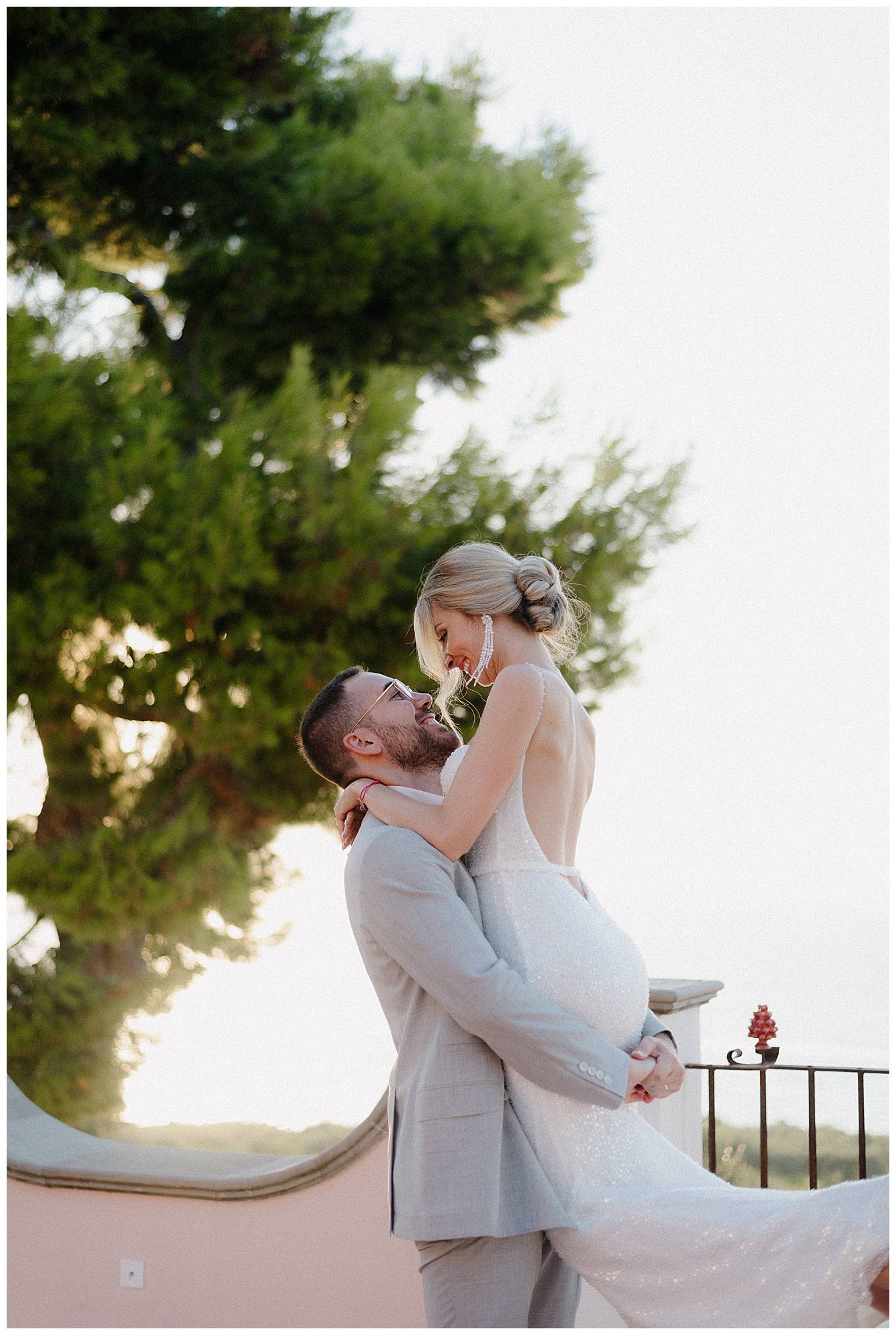 Romantic wedding moment captured as groom lifts bride in white dress against scenic outdoor background.