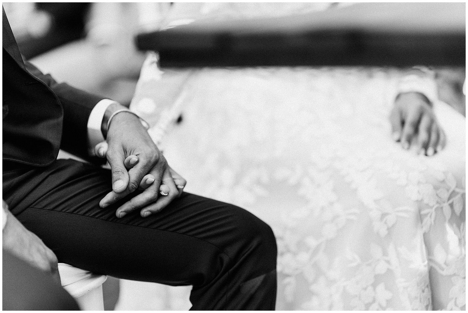 Black and white detail shot of newlyweds' hands resting on formal attire during wedding ceremony.