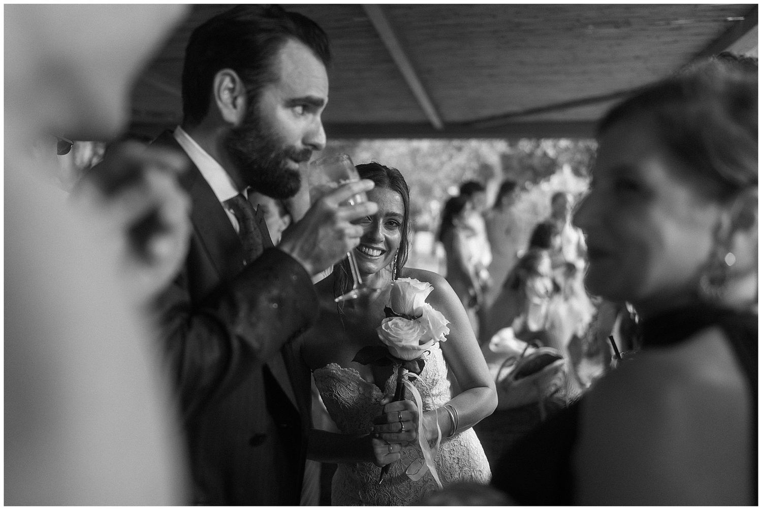 Black and white candid photograph of wedding guests sharing laughter and drinks during a celebration.