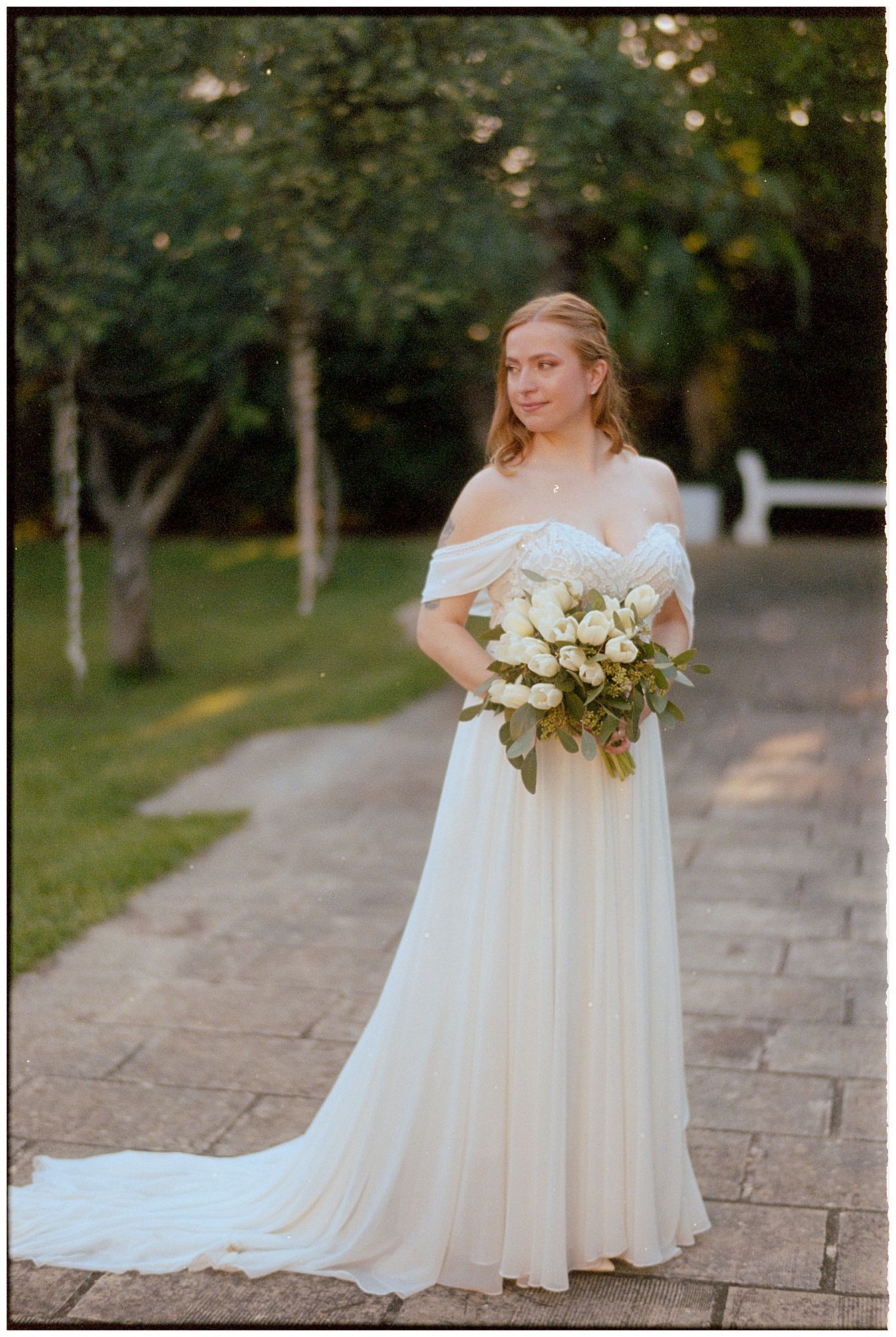 A person in a flowing off-shoulder white wedding gown holds a bouquet of white roses on a stone path in a garden setting.