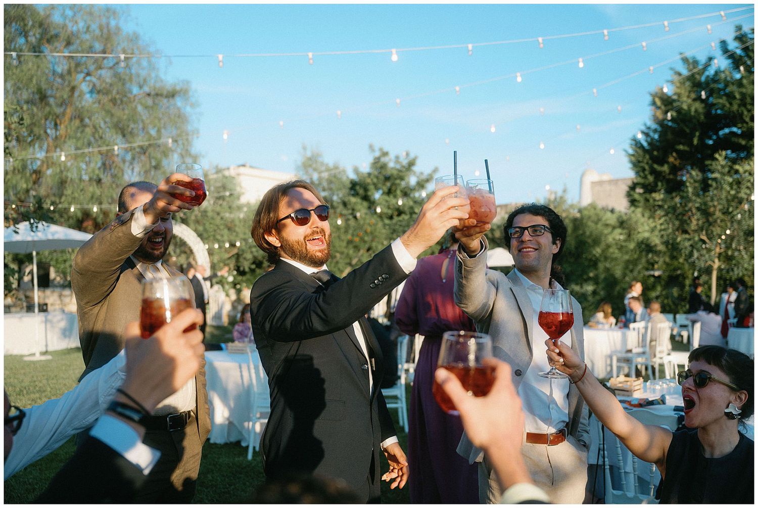 A group of young people raises their beverages in a celebratory toast outdoors on a sunny day.