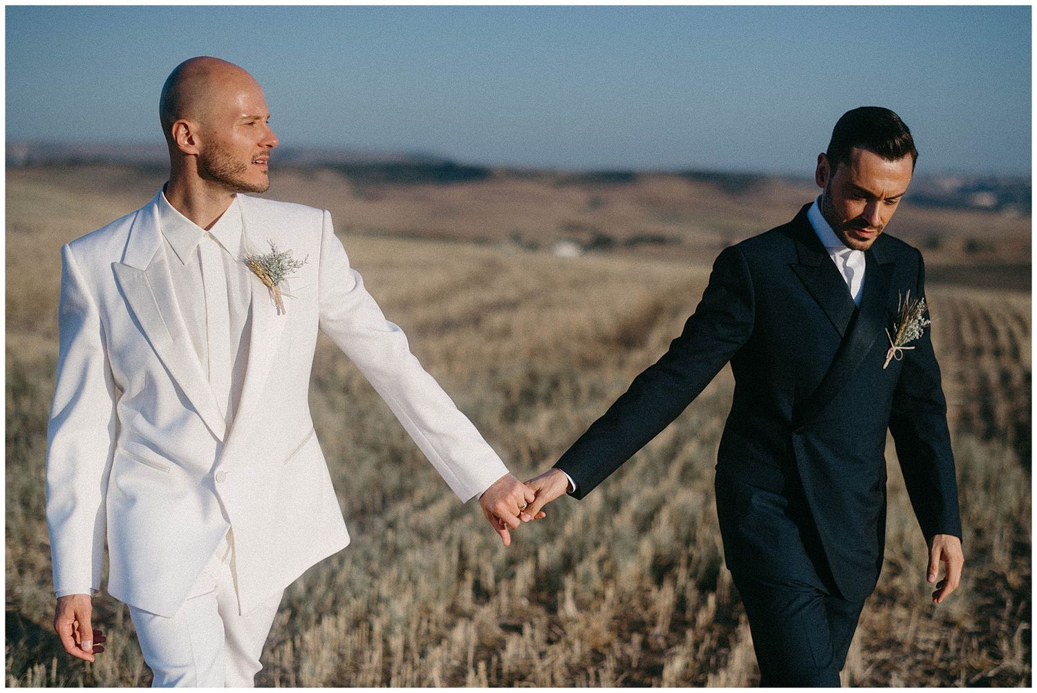 Two people in contrasting suits hold hands while walking through a scenic golden field at sunset.