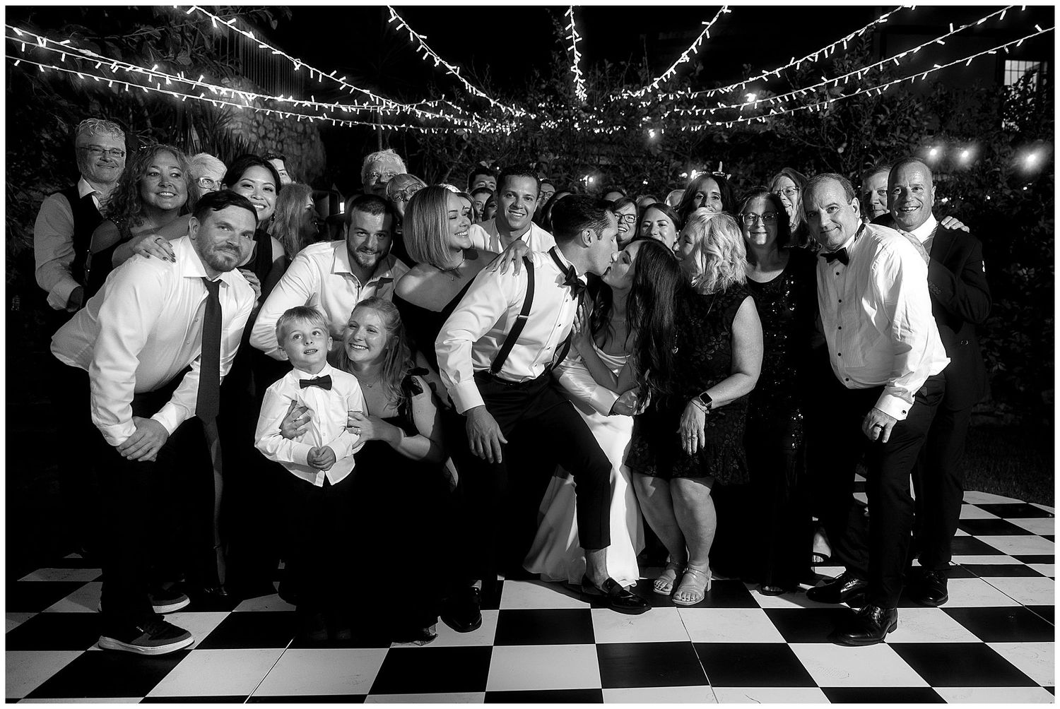 Black and white photo of party guests dancing and posing on a checkered dance floor under twinkling lights at night.