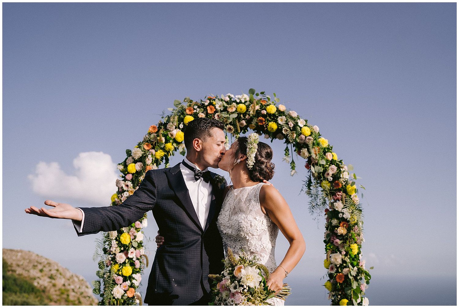Couple embraces under beautiful floral wedding arch with yellow and white flowers against scenic mountain backdrop.