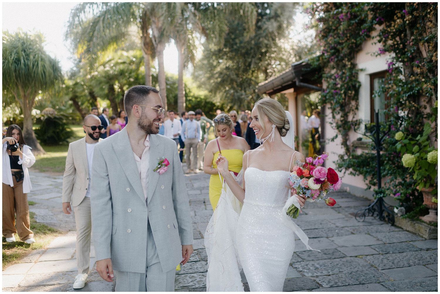 Newlywed couple walking outdoors at garden wedding venue with guests visible in background during sunset.