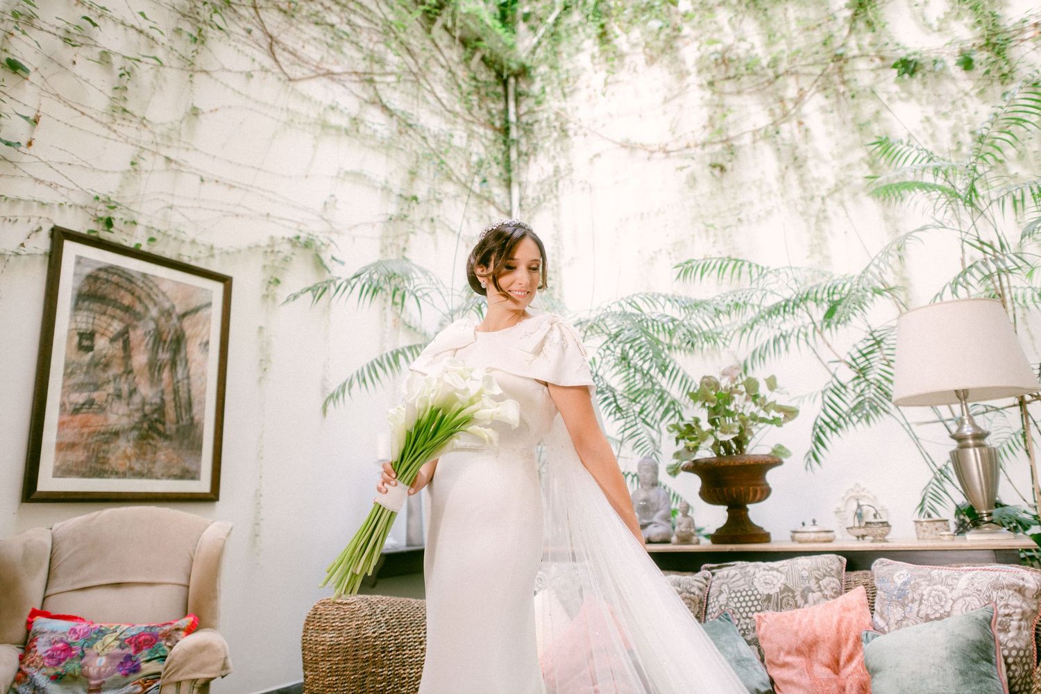 Elegant figure in flowing white gown poses in vintage-style room with lush greenery and framed artwork.