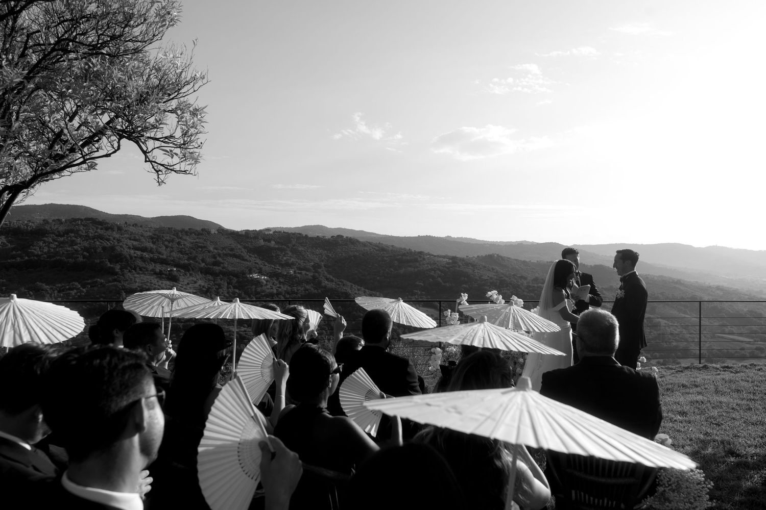 A silhouetted group of people holding white parasols stand on a hillside against a mountain landscape at sunset.