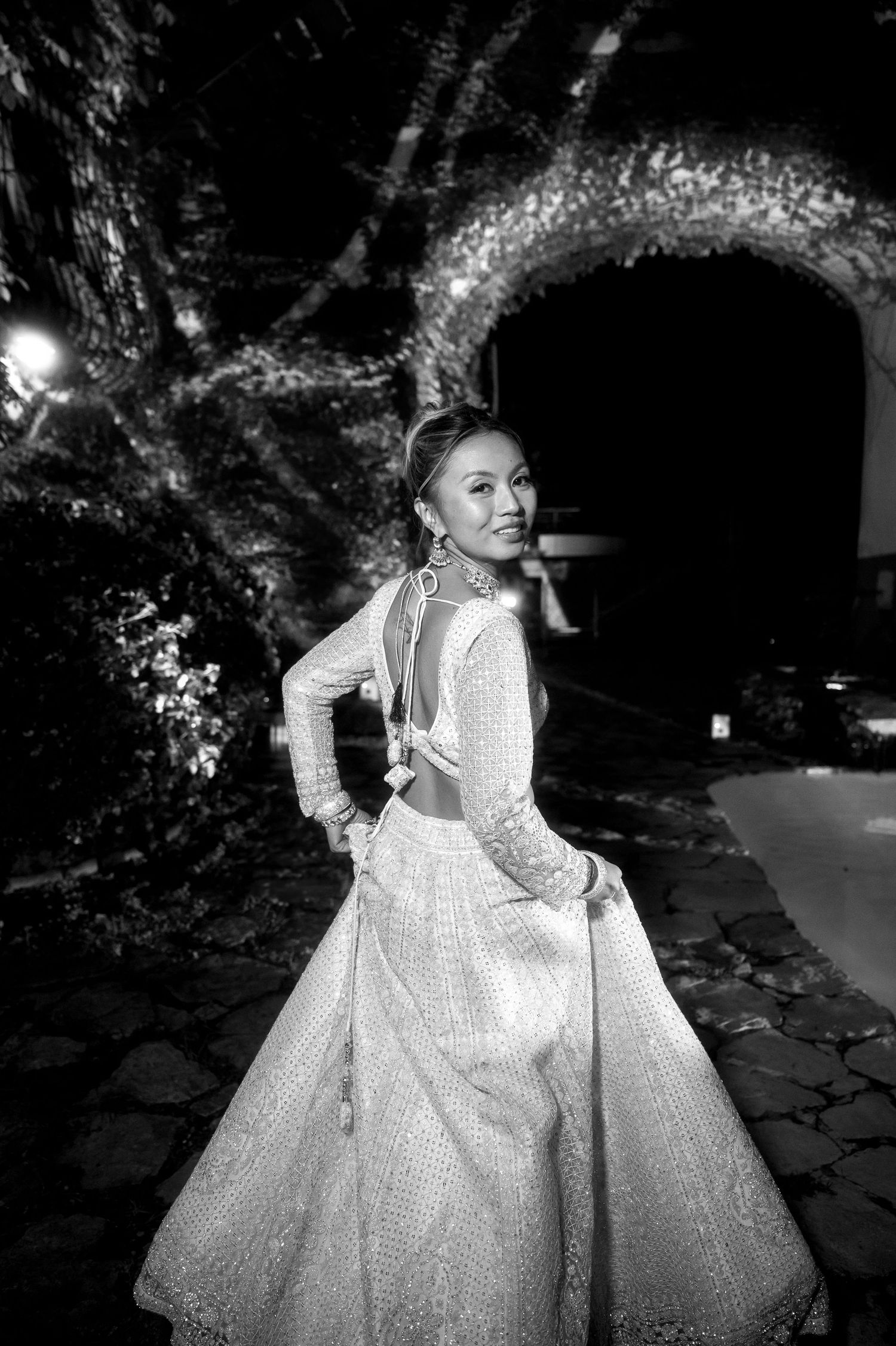 A bride in a sparkly white long-sleeved ballgown poses elegantly against a dramatic nighttime garden backdrop.