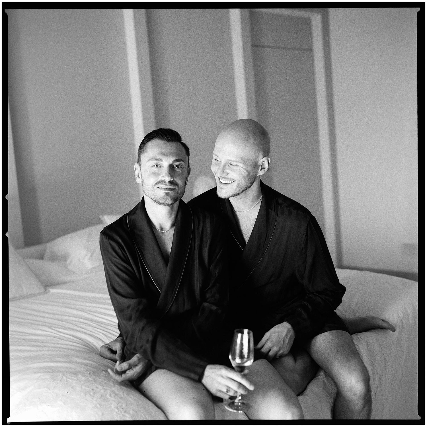 Two men in black clothing sit together on a white surface in a black and white photography series.