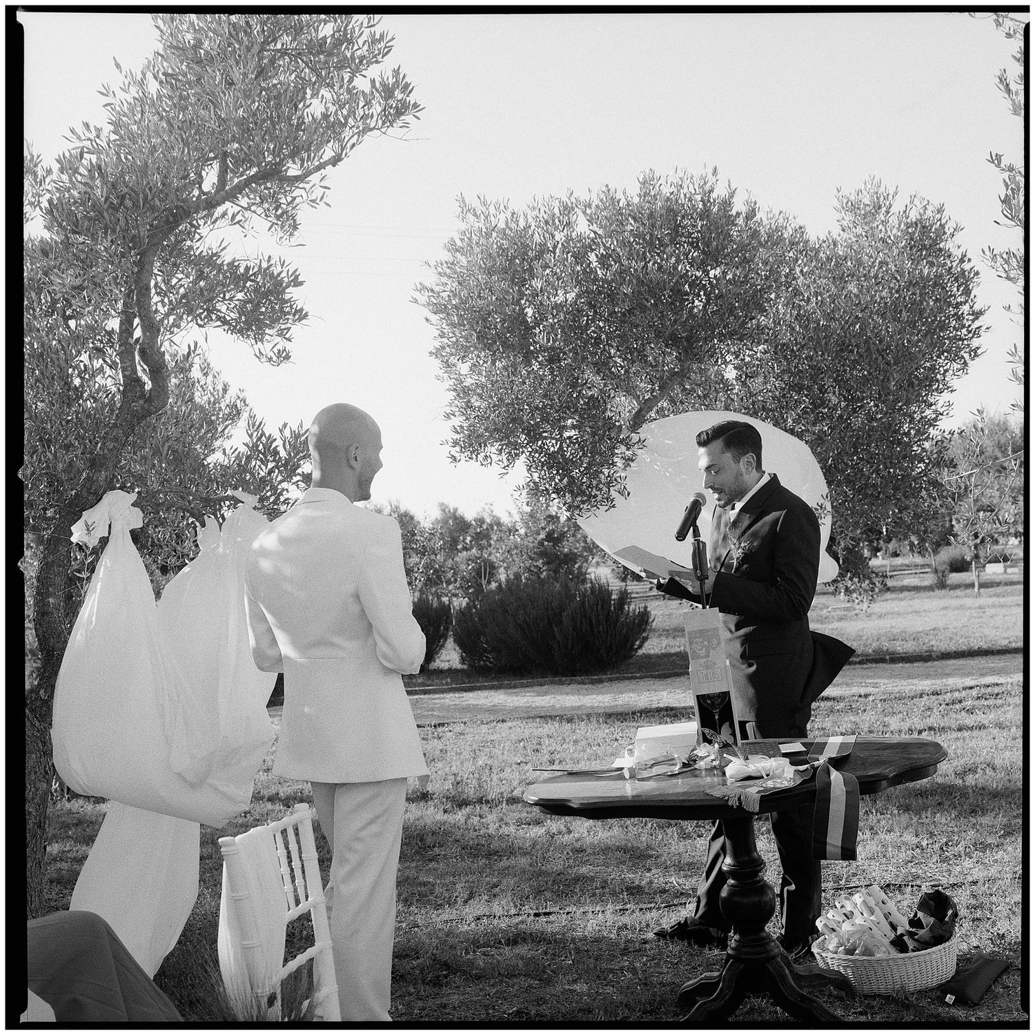 A black and white photograph shows a man standing on a bench playing guitar while a couple stands nearby in an outdoor setting.
