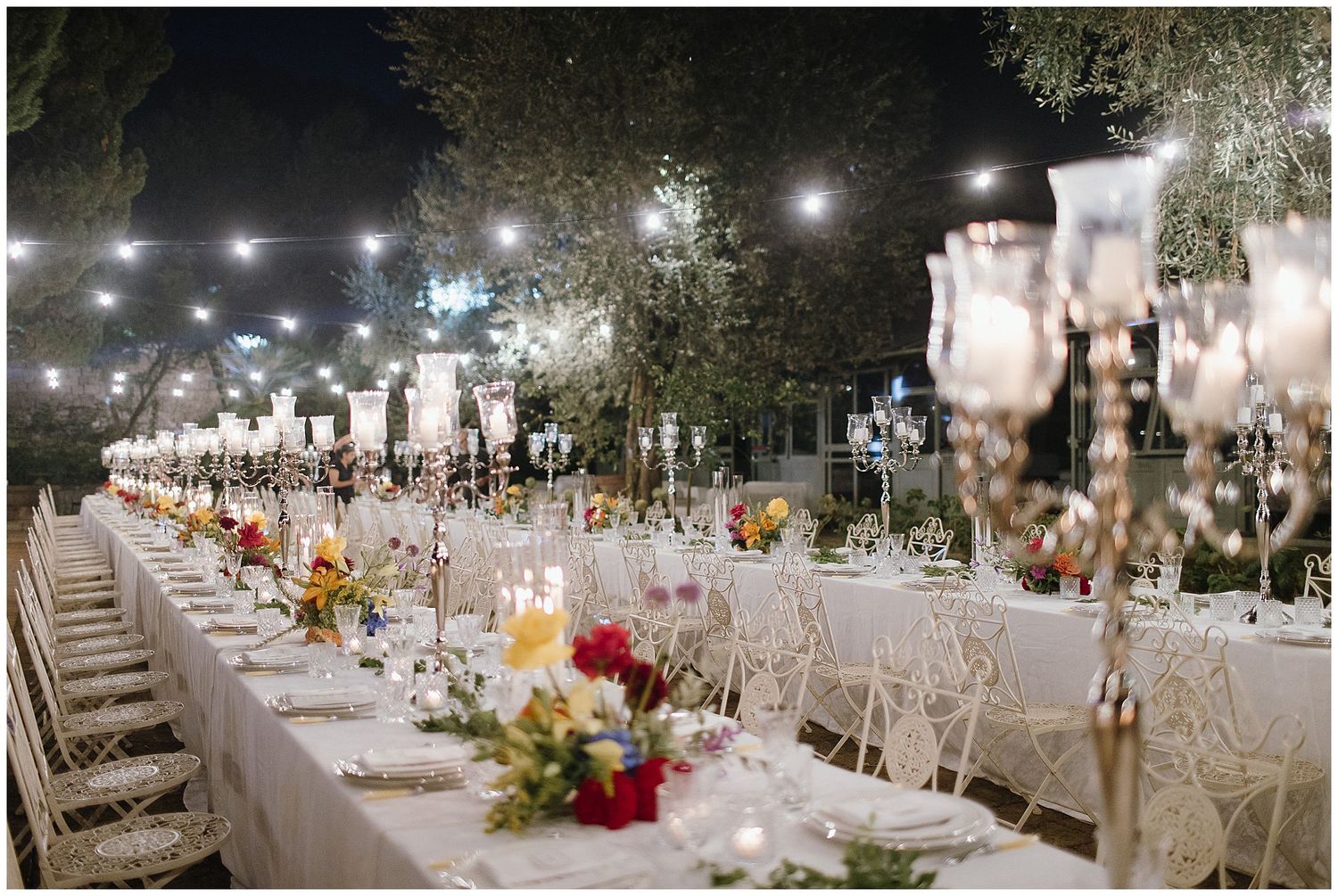 Elegant outdoor wedding reception table setting with long rows of white chairs and floral centerpieces under twinkling lights.