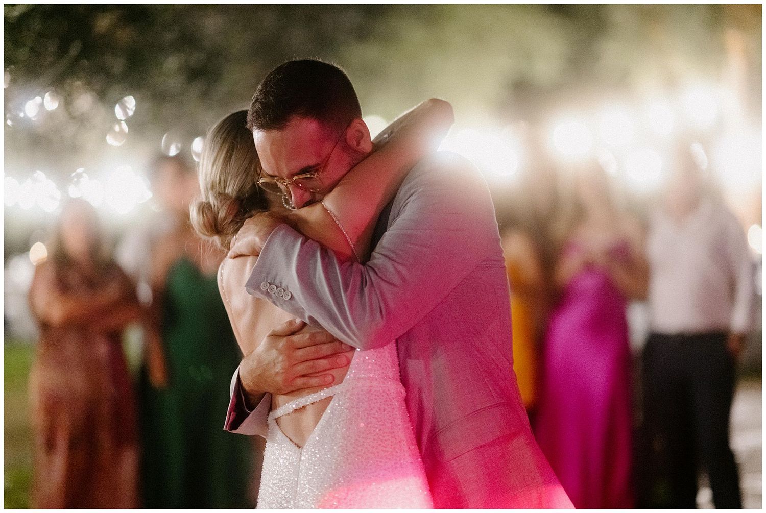 Newlywed couple shares intimate first dance at wedding reception with pink lighting and guests in background.