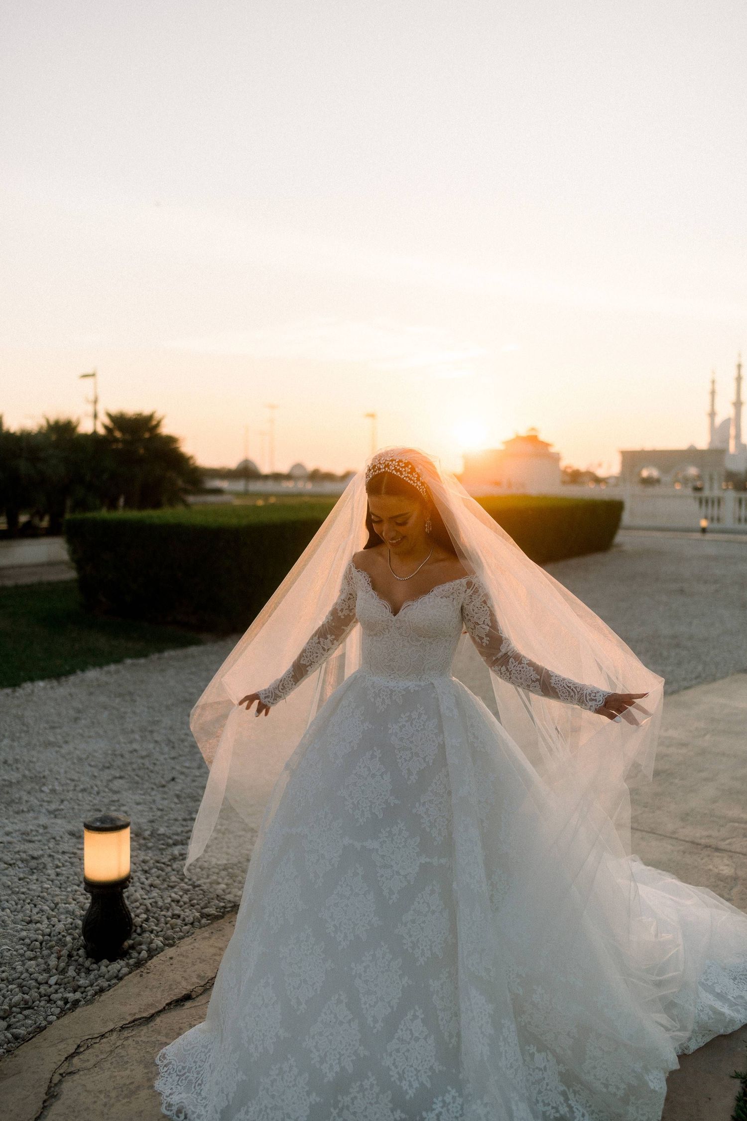 A bride in a white lace gown twirls her veil at sunset with a glowing lantern on the ground nearby.