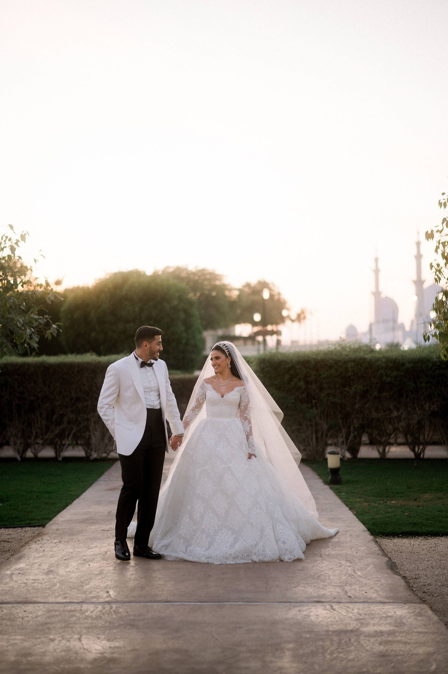 A couple in wedding attire walking along a garden path at sunset with distant architecture visible in the background.