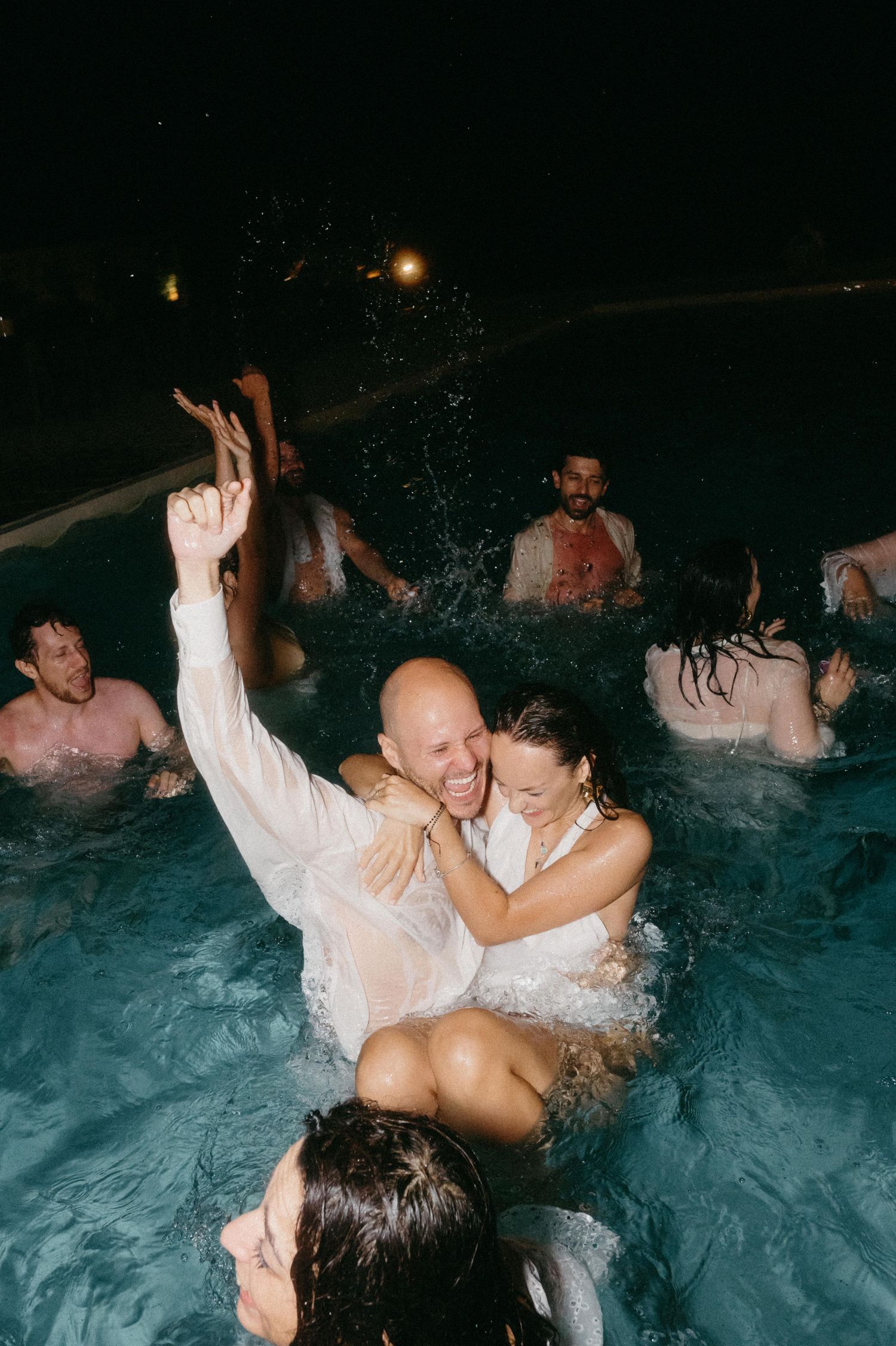 Wedding guests celebrate by jumping into a pool at night with splashing water and ambient lighting.