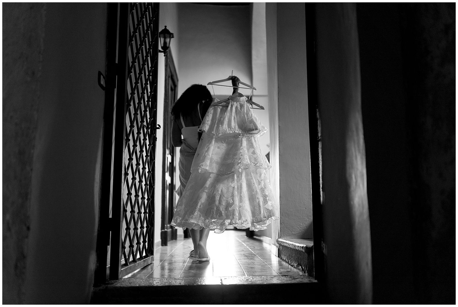 A wedding dress hangs in a doorway against backlit window, creating a dramatic black and white silhouette.