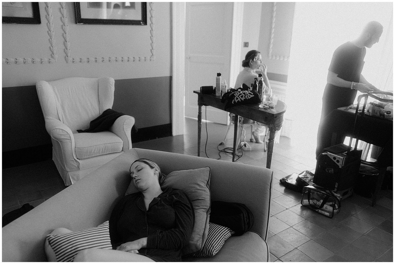 Black and white photograph showing a room with figures in various positions, including someone reclining on a couch.