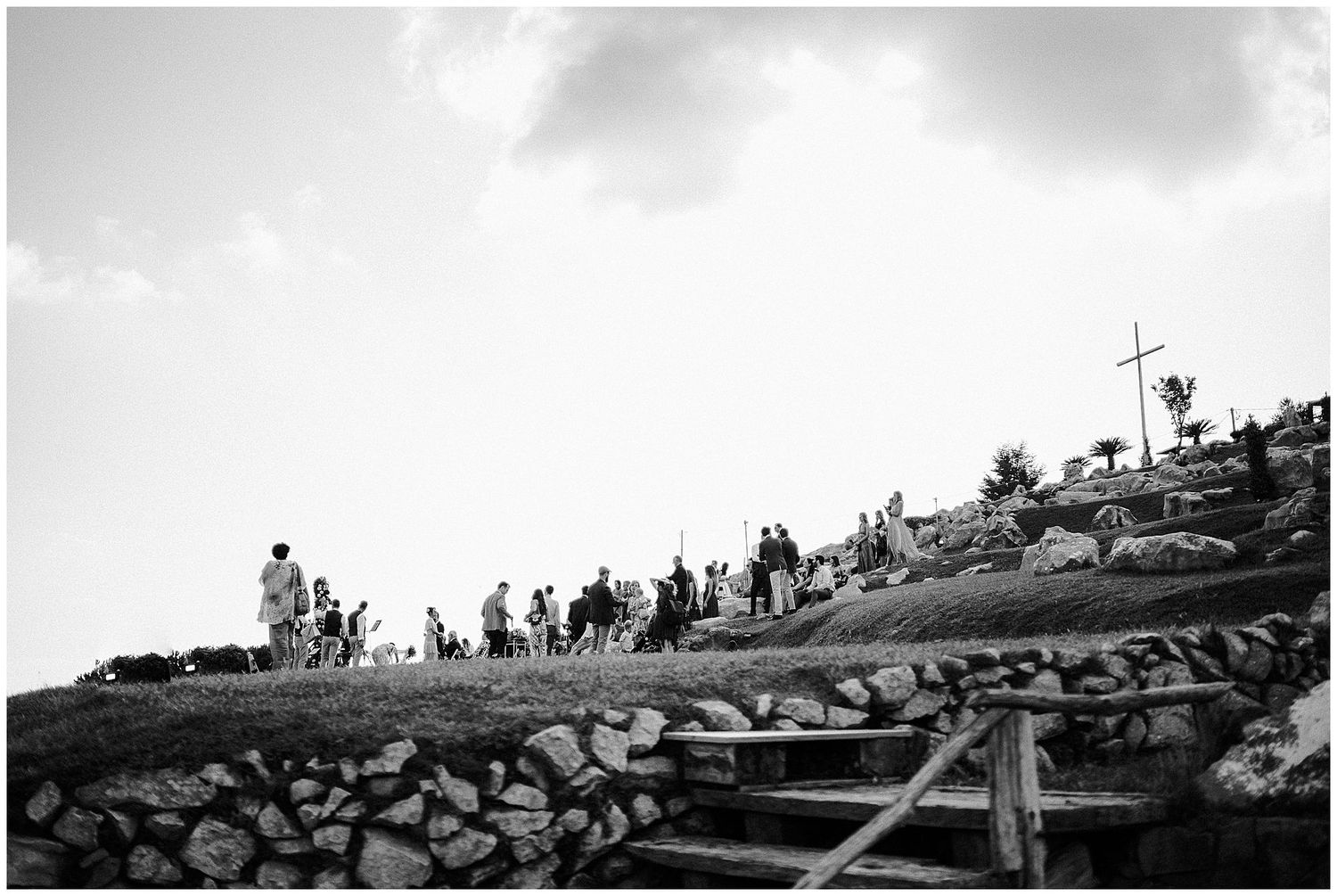Silhouetted pilgrims climb stone steps toward a hilltop cross in black and white photography.