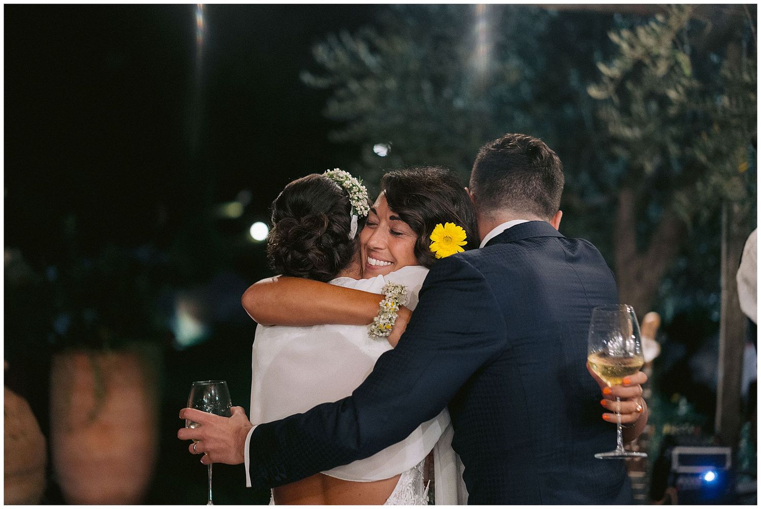Three friends embrace and share champagne during an outdoor evening celebration.