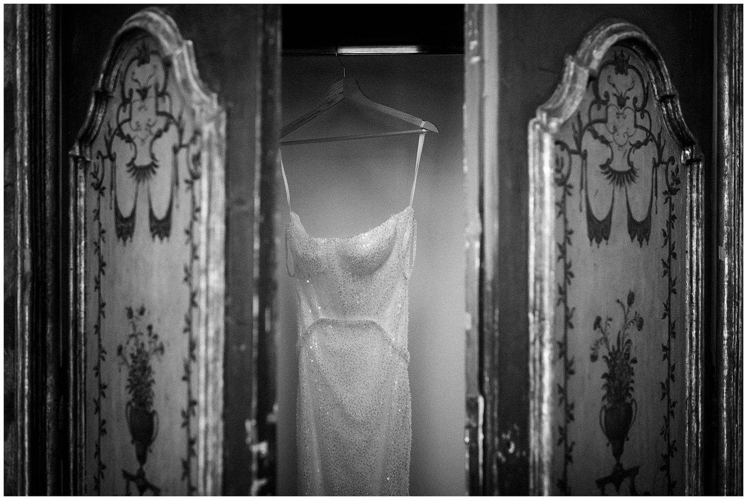 A white wedding dress hangs between ornate antique mirrored doors in a black and white photograph.