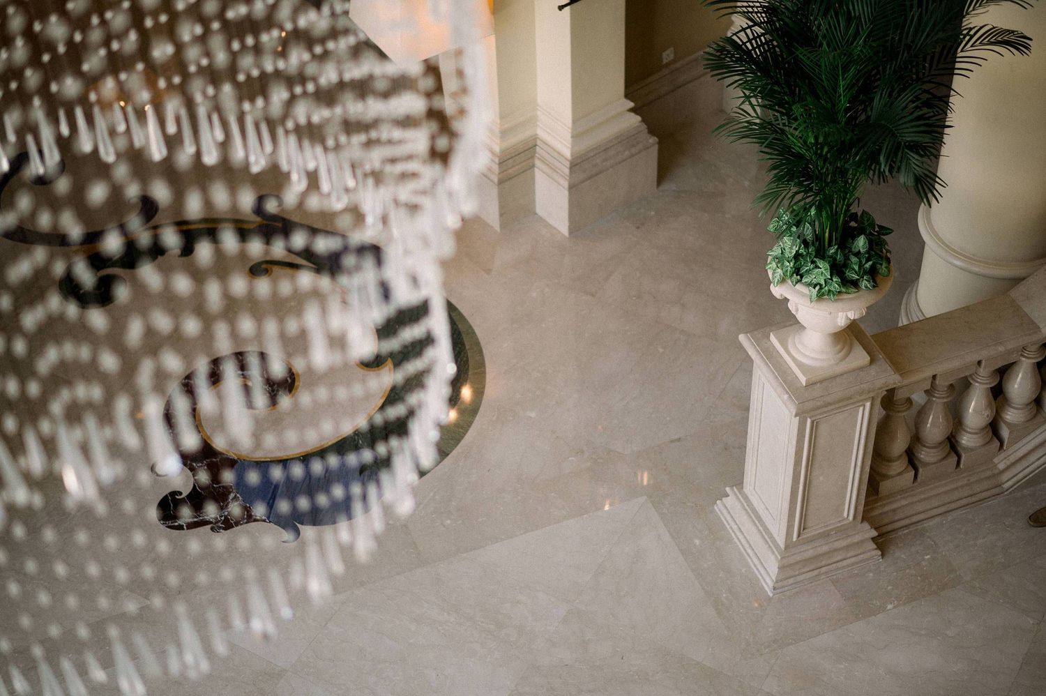 A crystal chandelier hangs above marble floors next to an ornate banister and decorative potted plant in an elegant space.