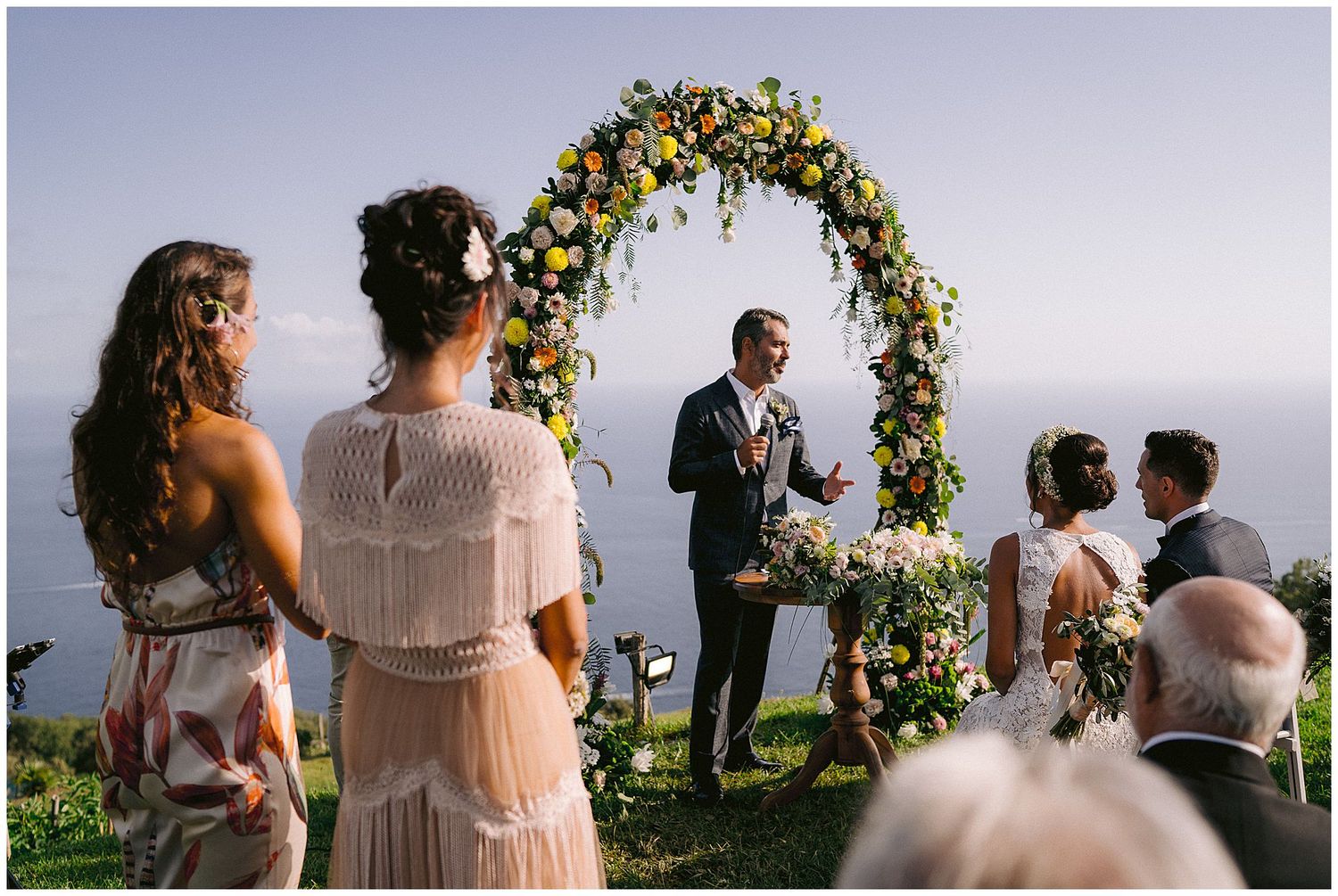 An elegant floral wedding arch decorated with yellow and white flowers stands overlooking a scenic outdoor ceremony.
