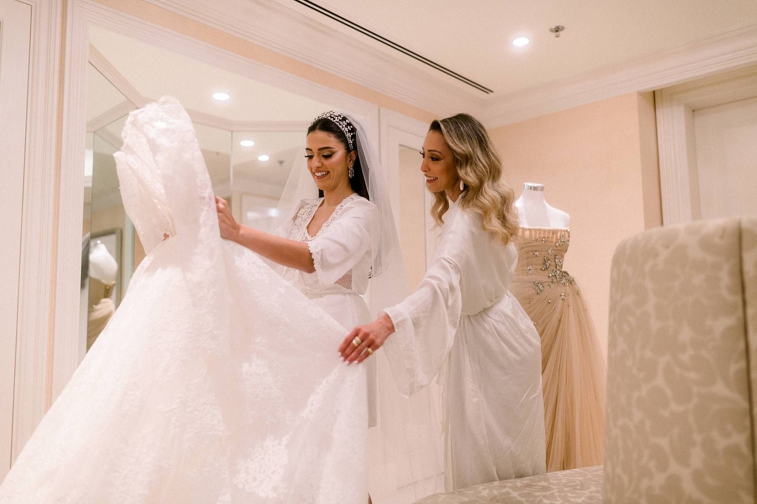 Two people in white robes examining an elegant white wedding dress in a bridal boutique.