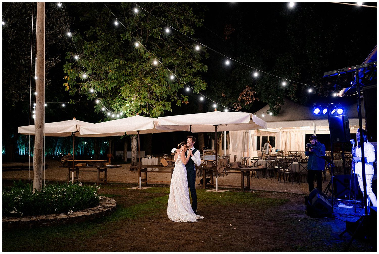 Newlyweds share an intimate dance moment under string lights at an outdoor evening venue.