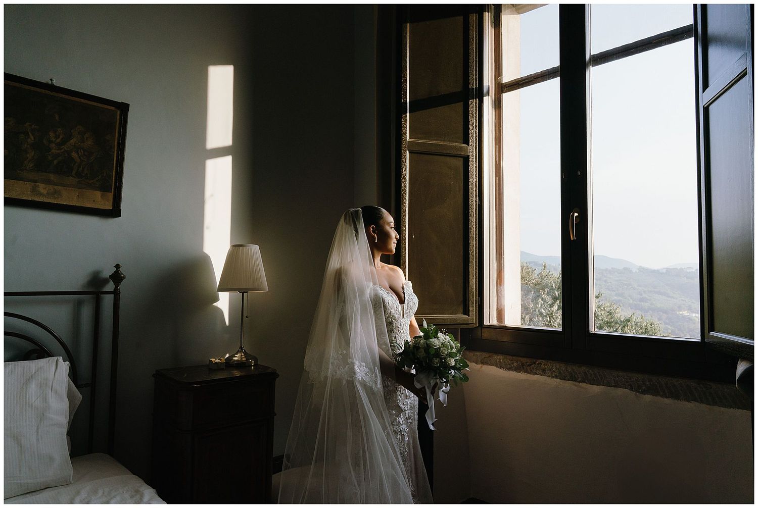 A bride in white dress and veil stands near window with bouquet in a romantic bedroom setting.