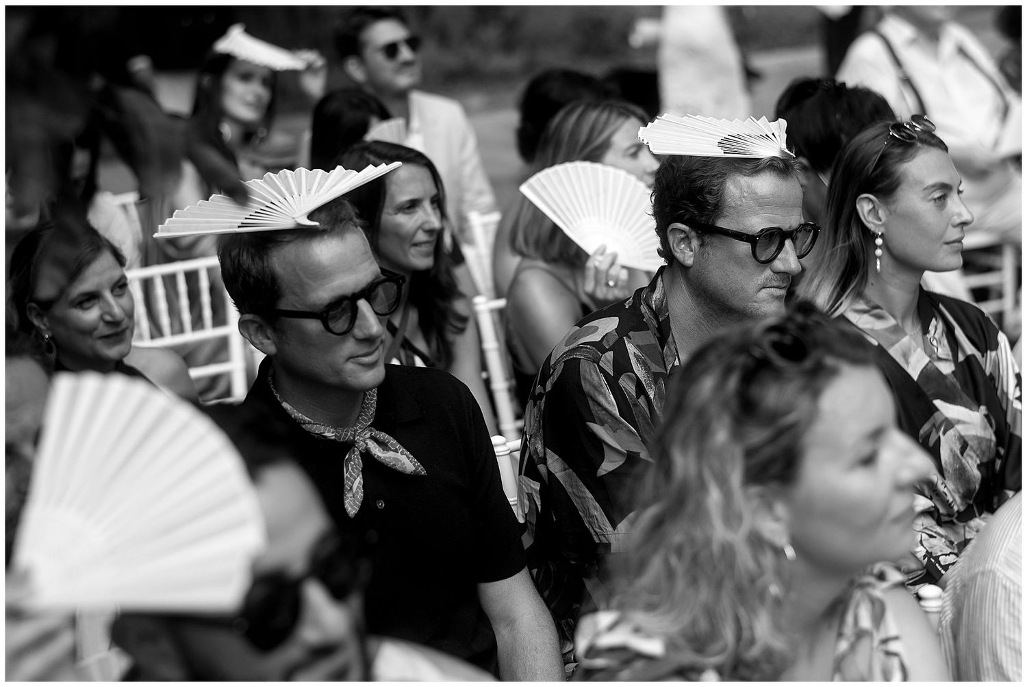 Vintage black and white photo of wedding guests with paper fans sitting in formal attire during an outdoor ceremony.