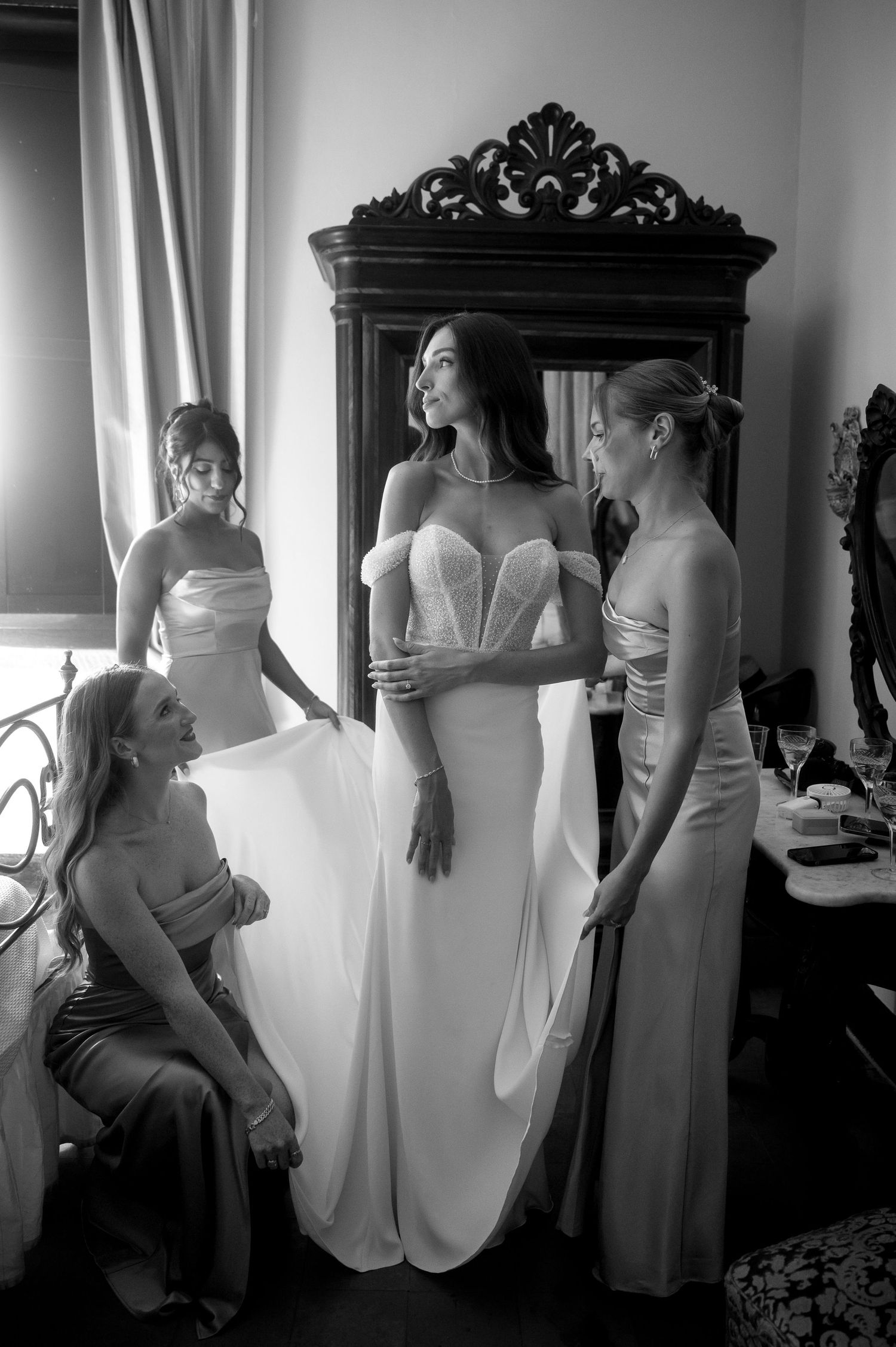 A bridal party gets ready in an elegant room with an ornate mirror, showing bridesmaids in light-colored dresses.