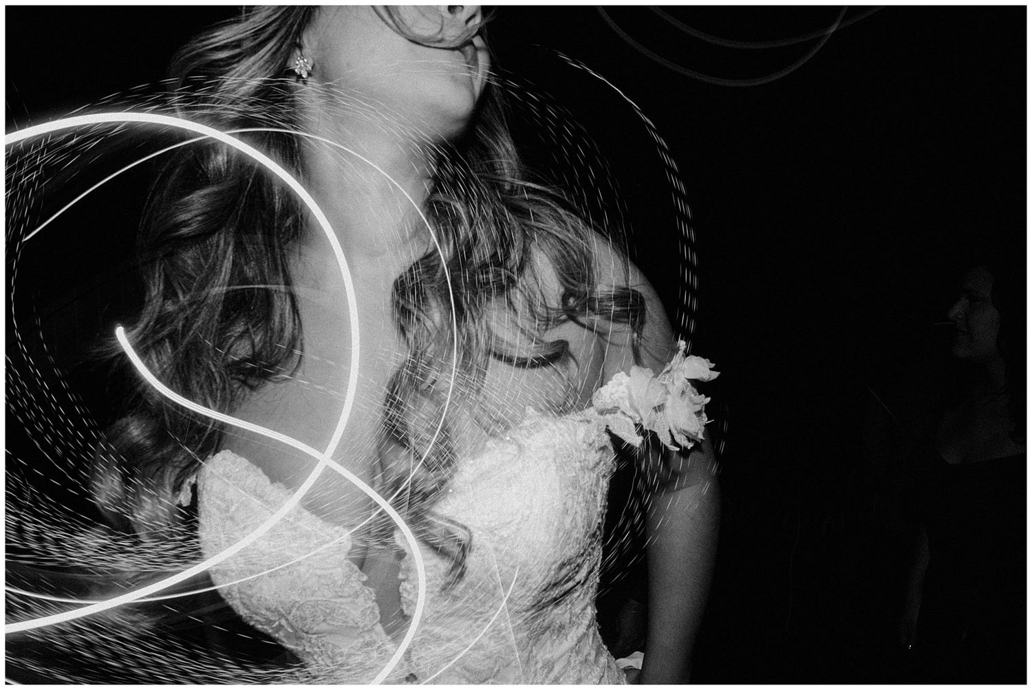 Black and white photo showing dramatic light trails and movement against a dark background with lace dress details visible.