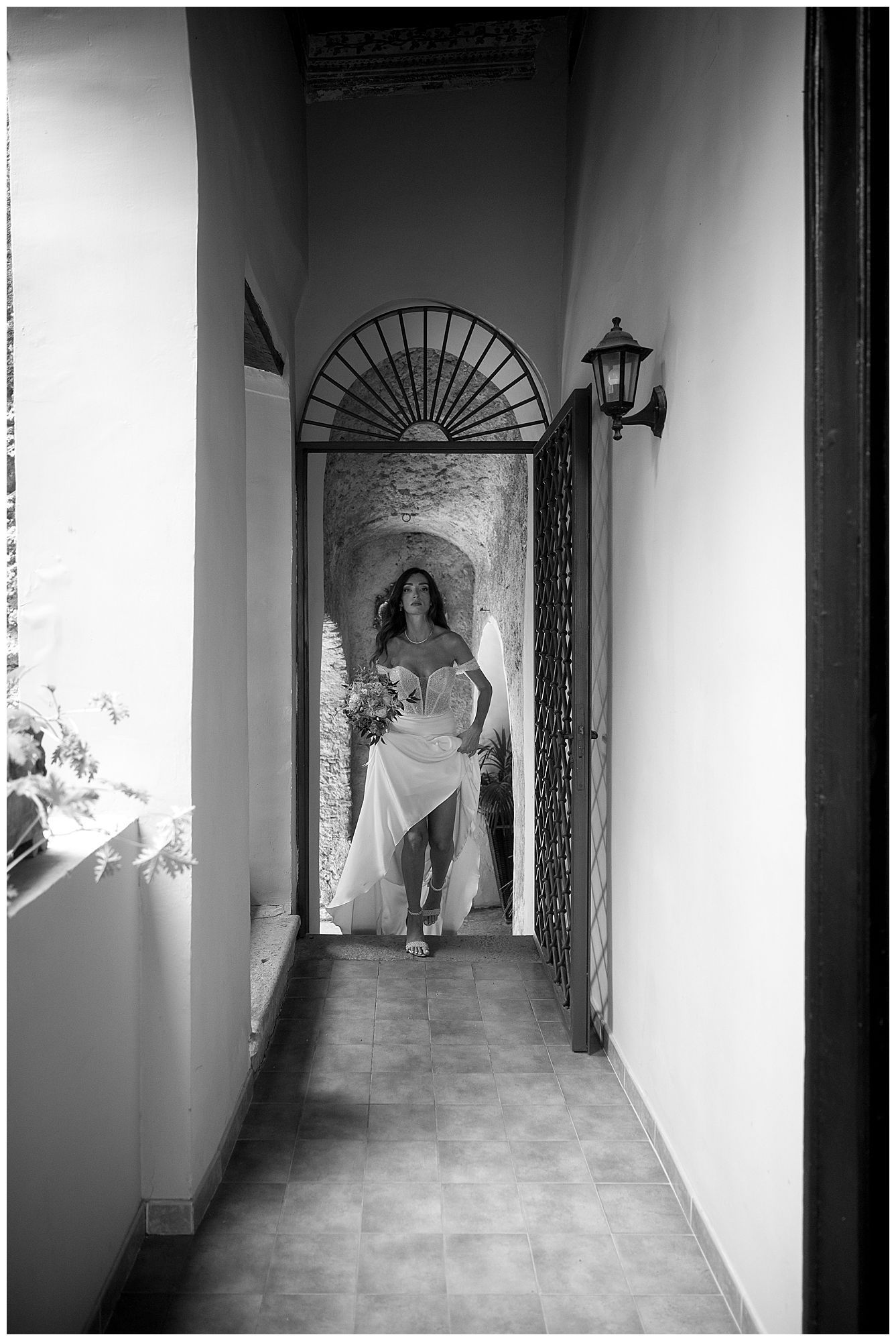 A black and white photograph shows someone in a white dress standing in an elegant hallway with arched doorway and lantern.