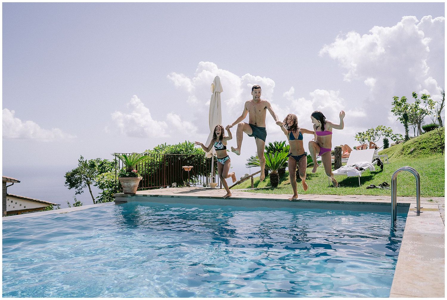 Group of people jumping into a luxurious infinity pool overlooking a scenic landscape on a bright sunny day.