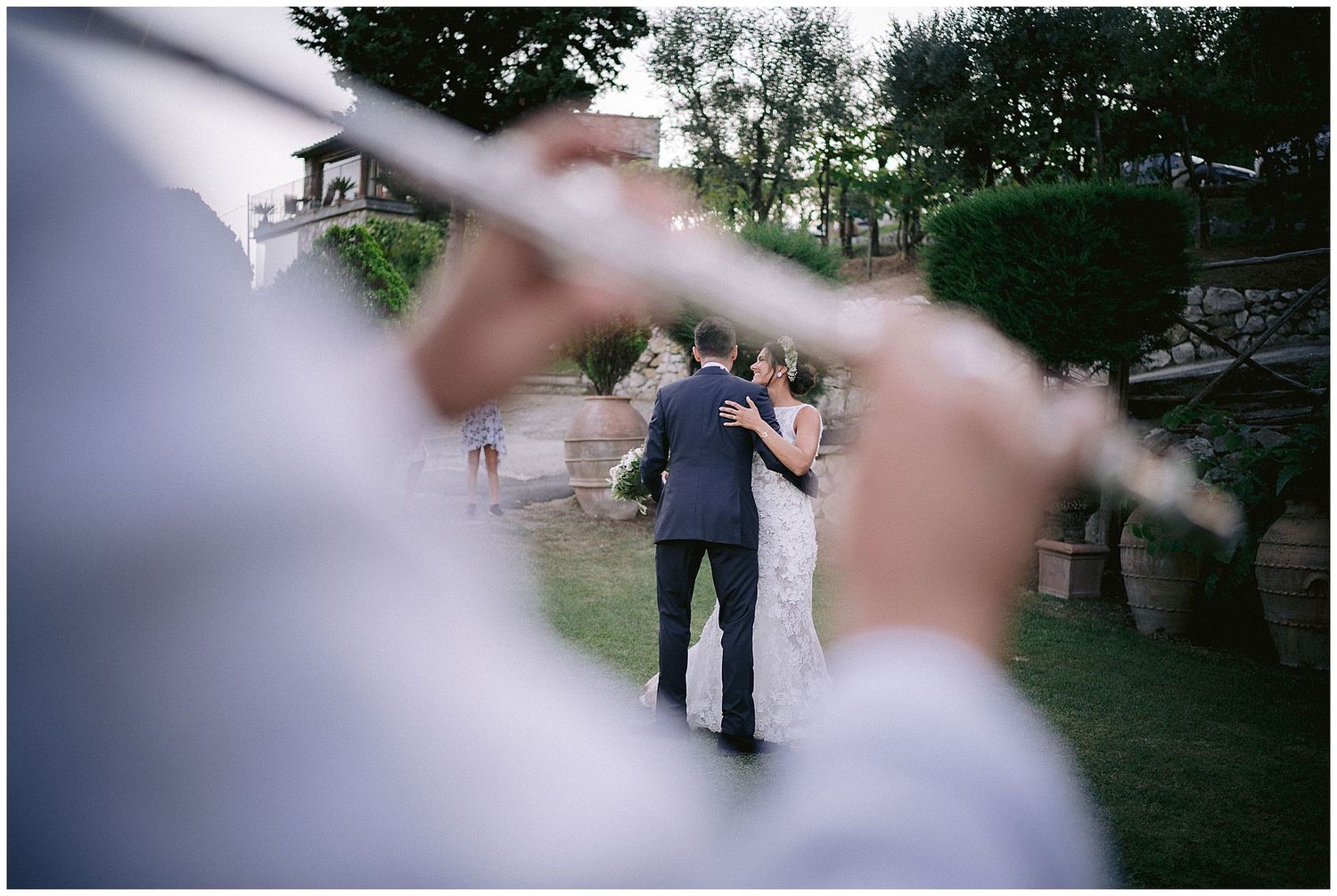 A couple dances at an outdoor wedding reception, viewed through blurred foreground elements in a candid photo.