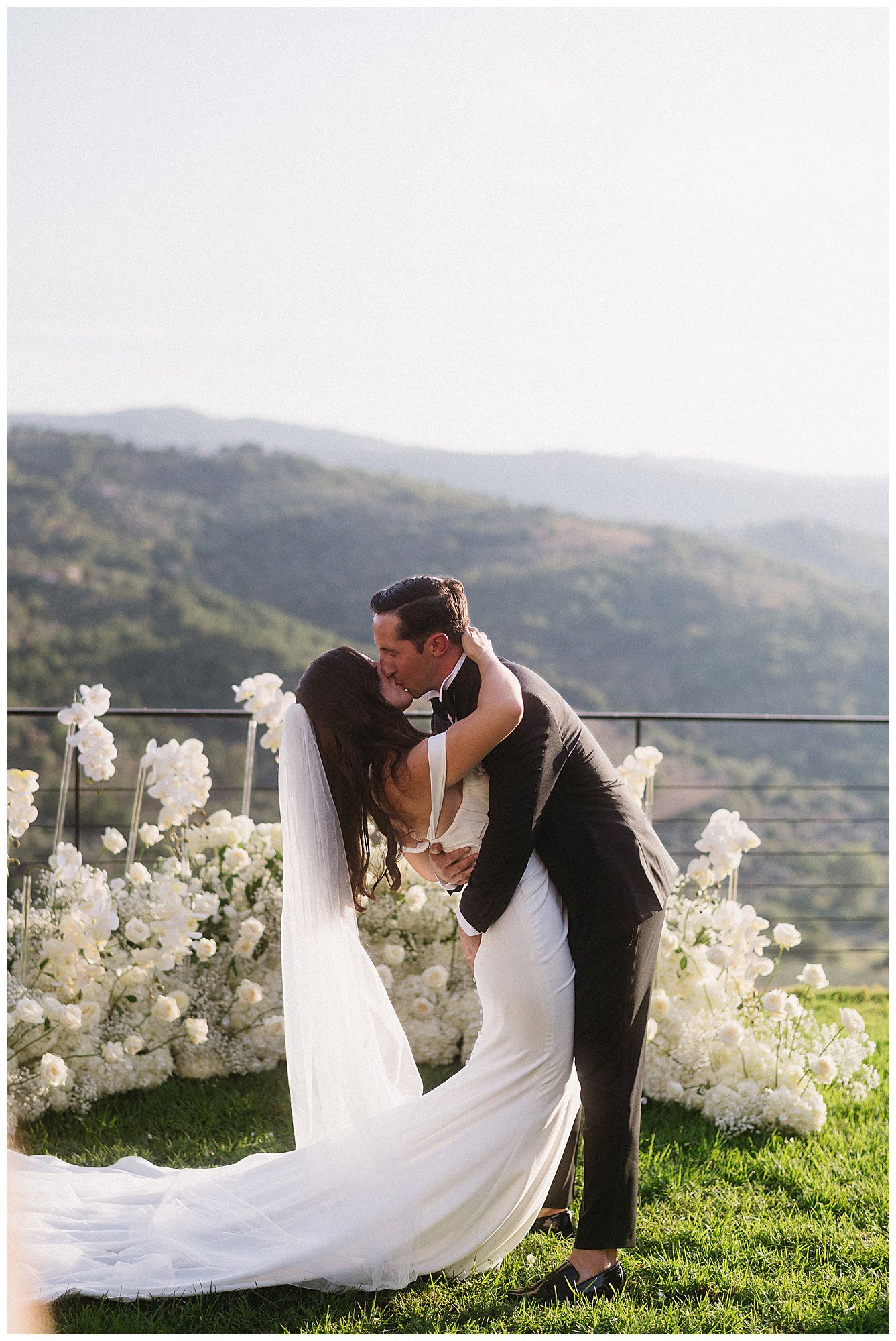 Romantic wedding moment photographed against mountain backdrop with white floral decorations on grassy terrace.