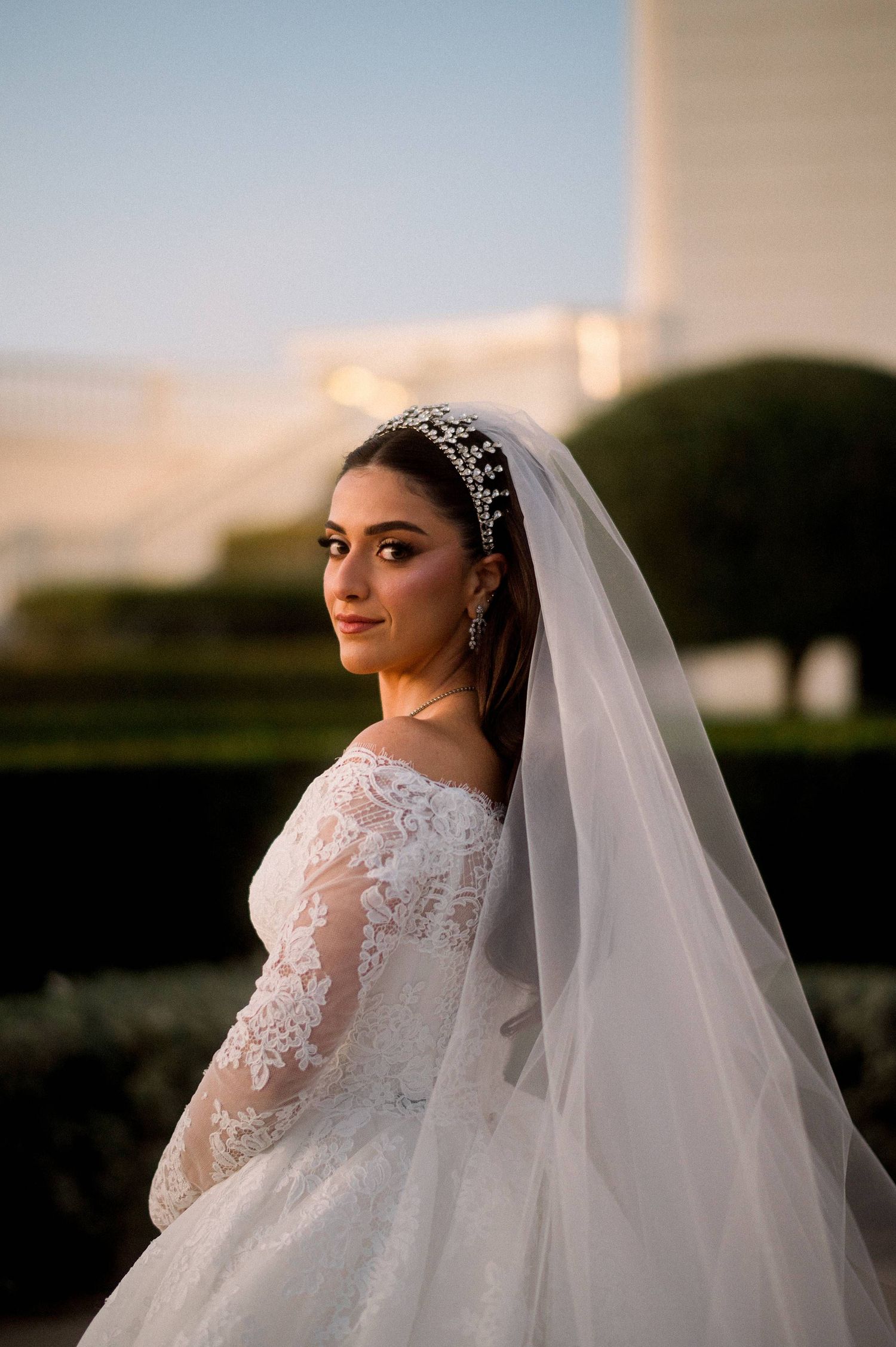 A bride in a long sleeve lace wedding dress with a crystal headpiece and flowing veil poses at sunset.
