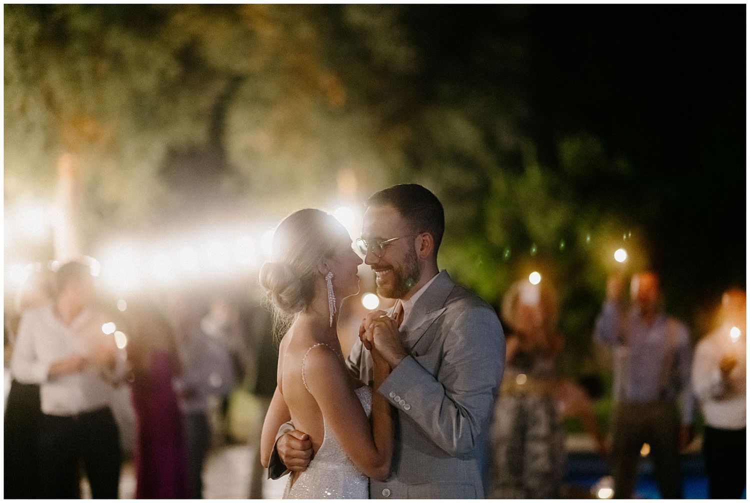 A couple shares their first dance at an outdoor evening wedding reception with soft lighting and guests in the background.