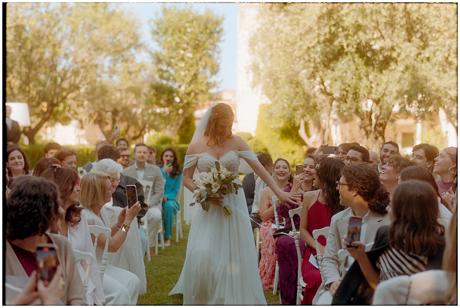 A joyful outdoor wedding celebration with guests gathered on a lawn during golden hour as a bride walks through the crowd.
