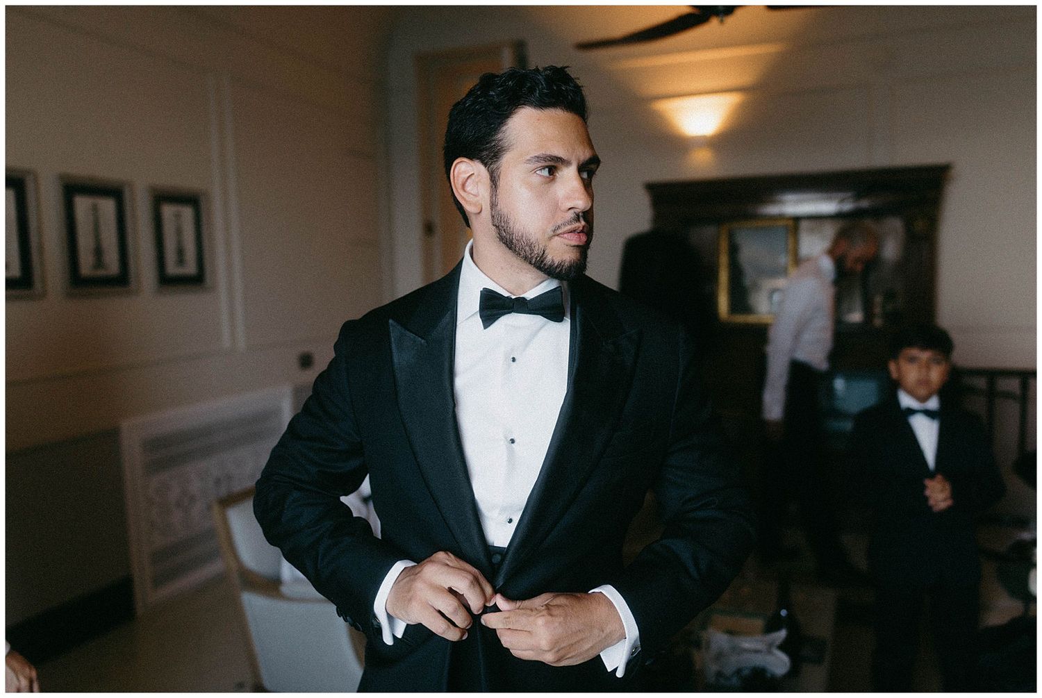 A person in a black tuxedo and bow tie adjusts their jacket in an indoor setting with framed artwork on the walls.