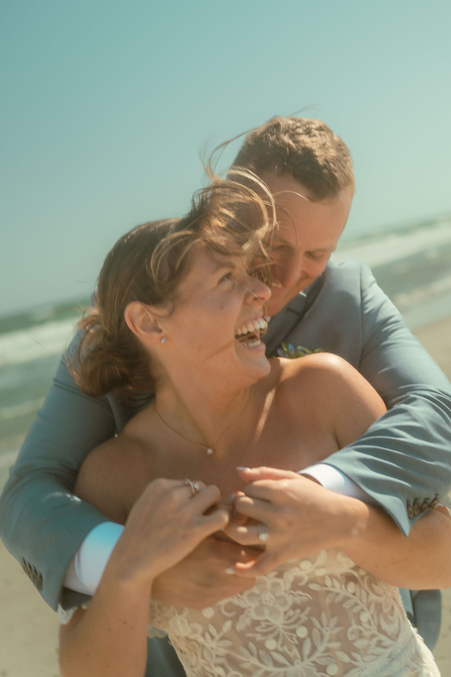 Joyful wedding couple embracing on a sunny beach with bride wearing lace dress.