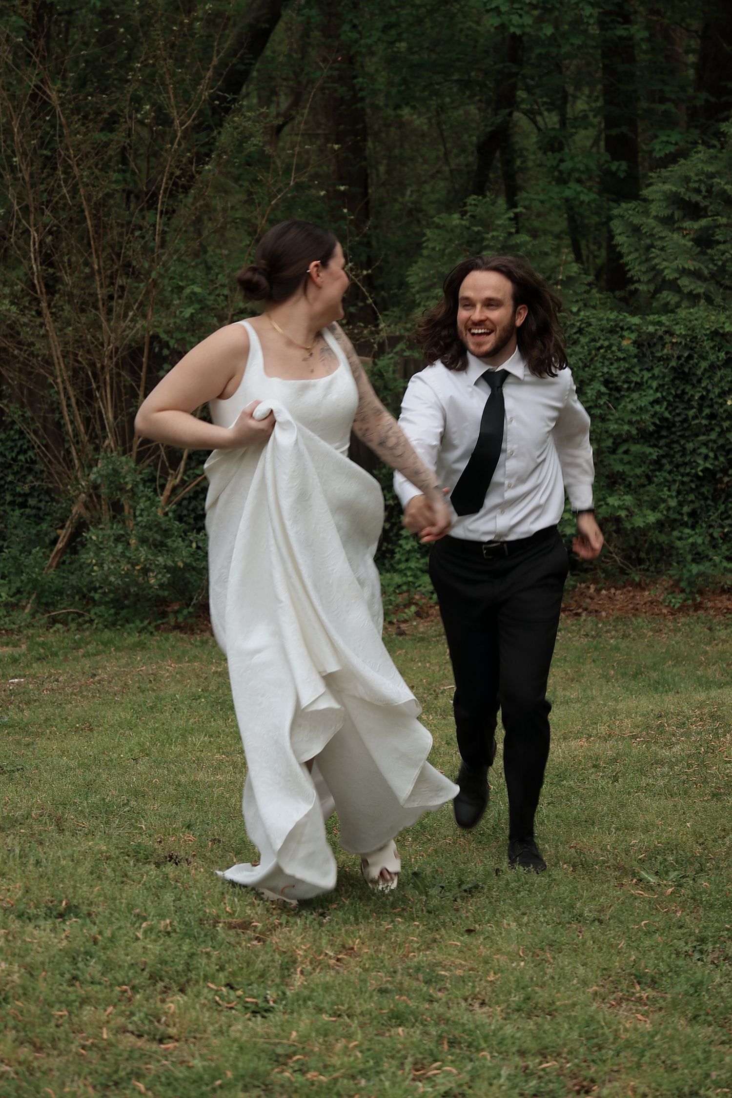 Happy couple running through wooded area, bride in flowing white dress and partner in formal attire.
