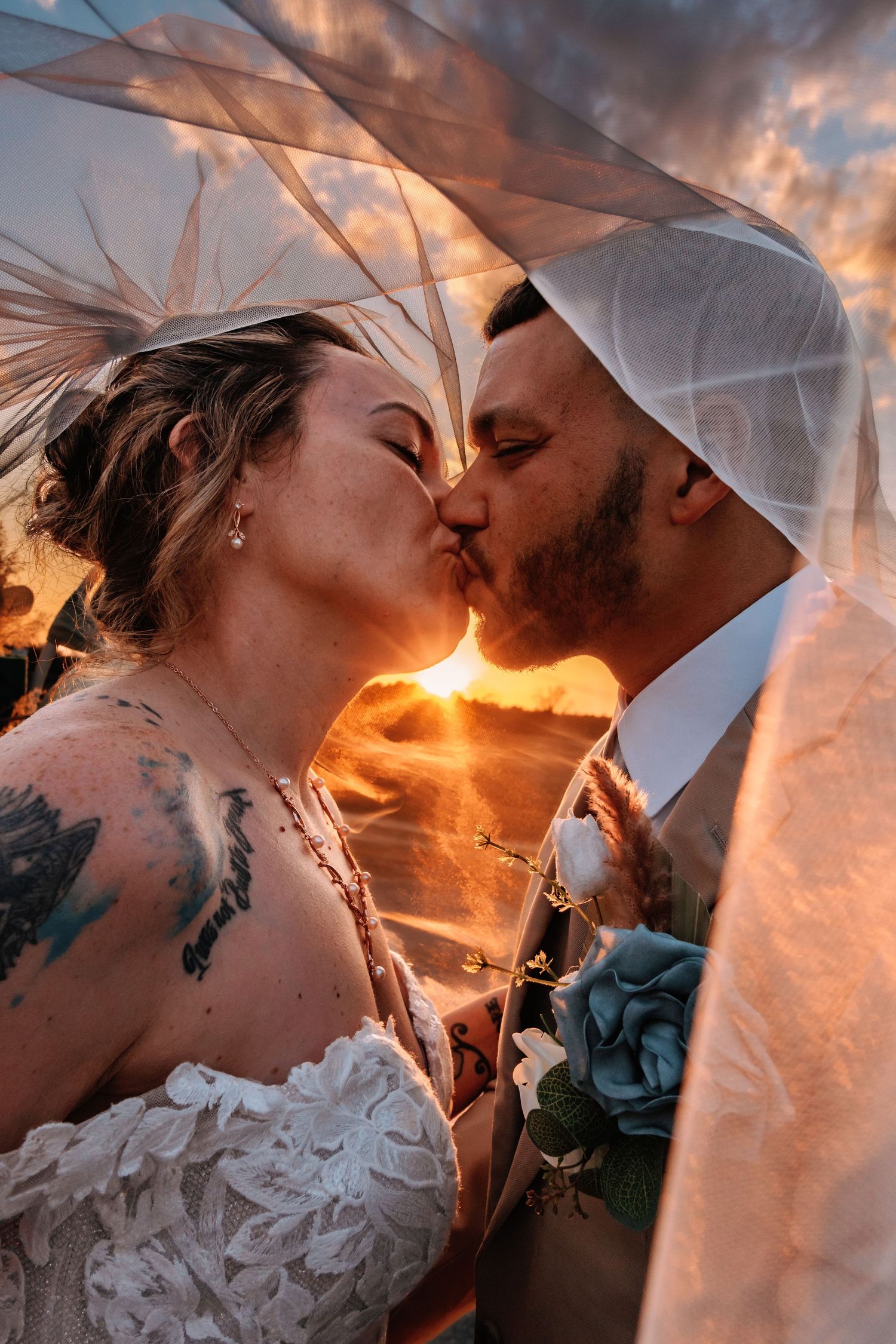 Romantic wedding kiss at sunset with flowing veil creating ethereal atmosphere against orange sky.