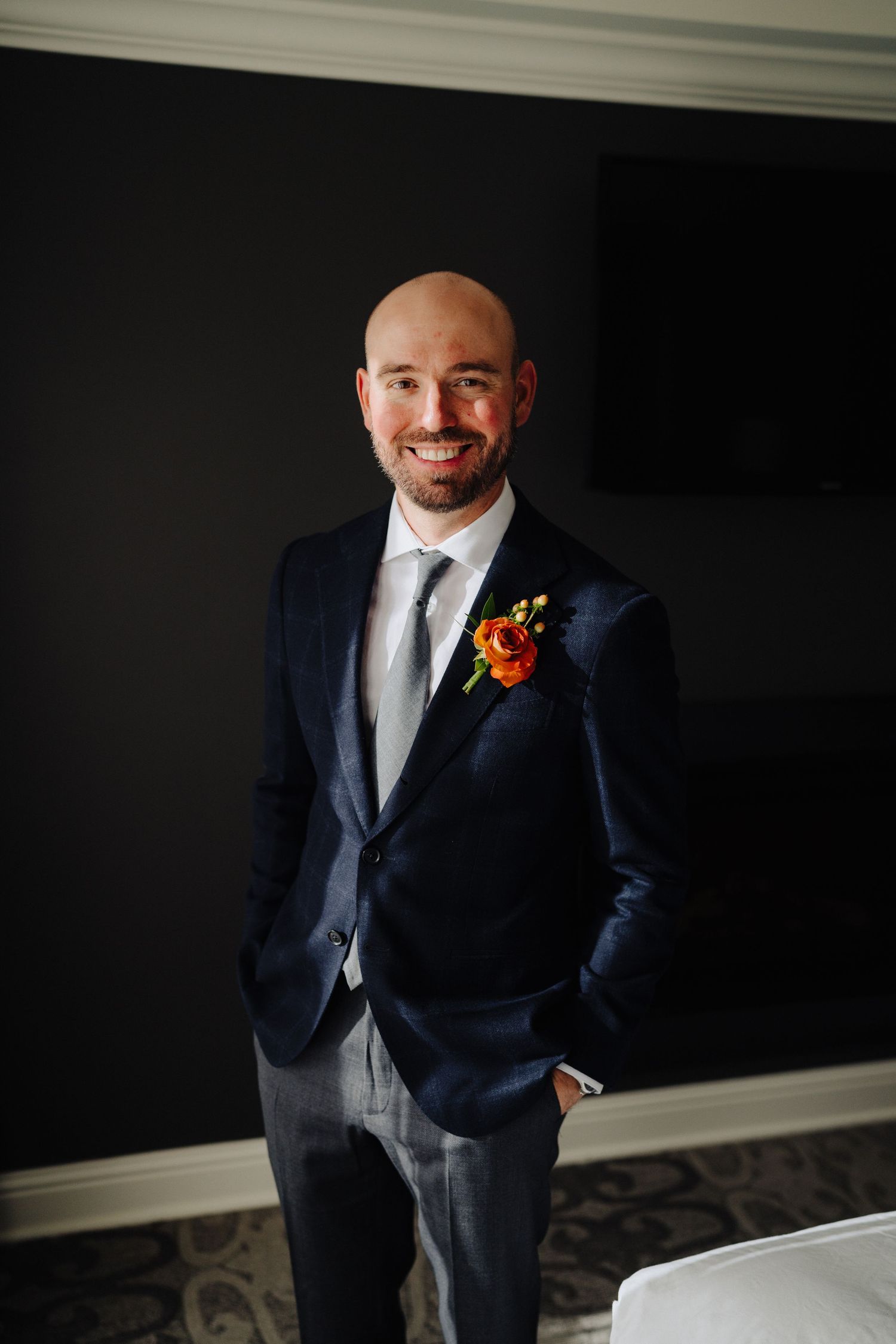 A groom wearing a navy suit and orange boutonniere poses confidently against a dark background.