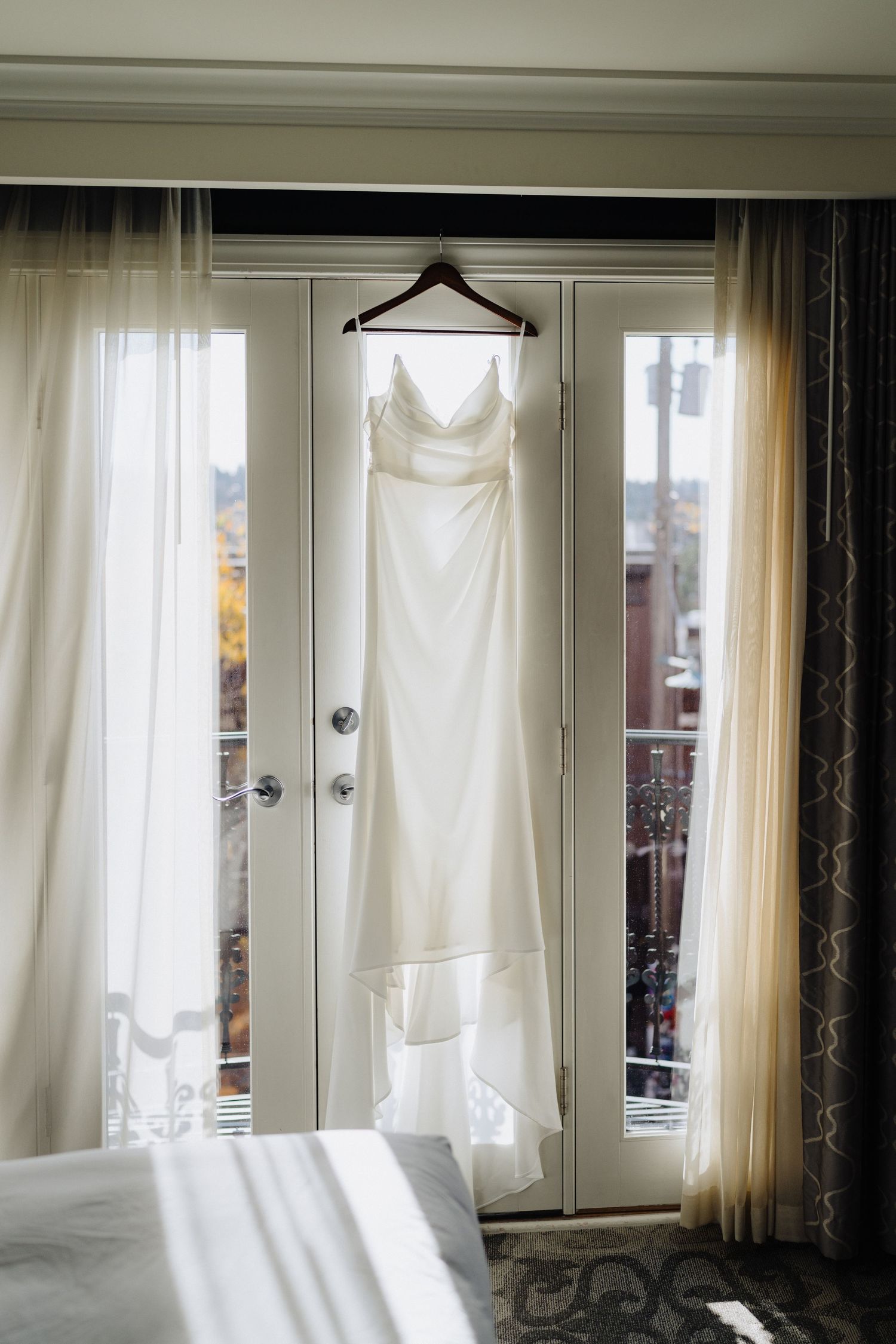 An elegant white wedding dress hanging on a wooden hanger against French doors with sheer curtains in natural light.
