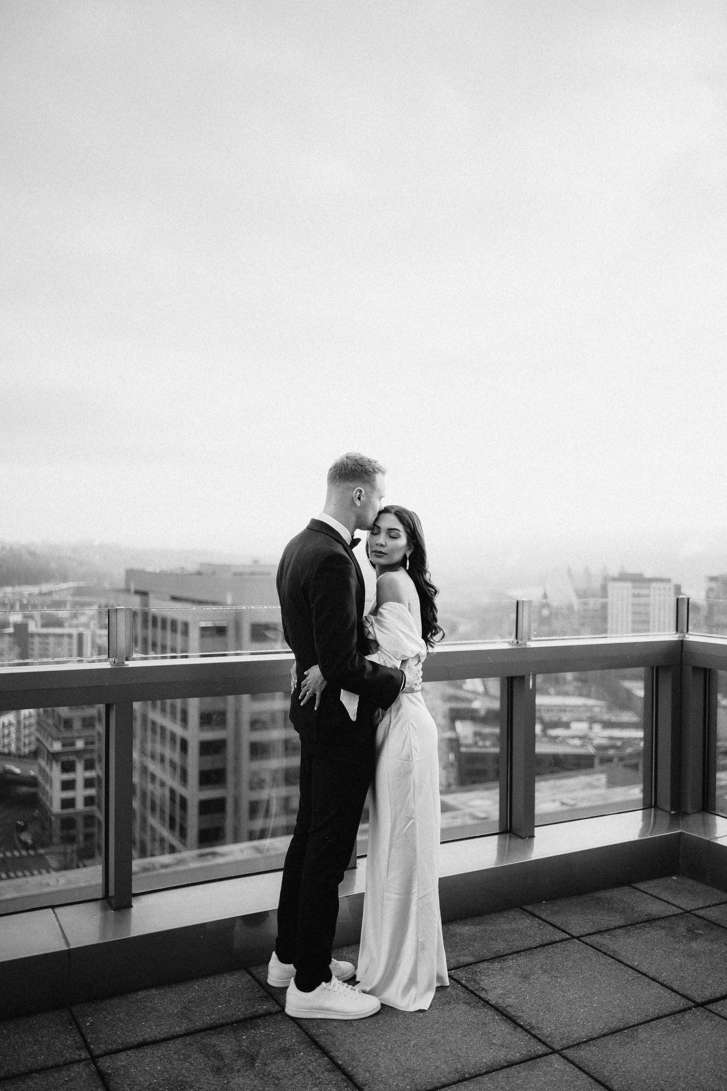 A couple shares an intimate embrace on a rooftop balcony overlooking a cityscape in black and white.