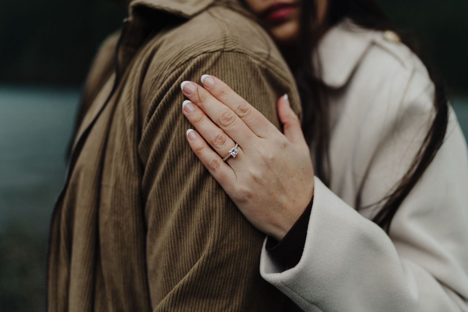 Close-up of engagement ring on hand against brown jacket with lake in background.
