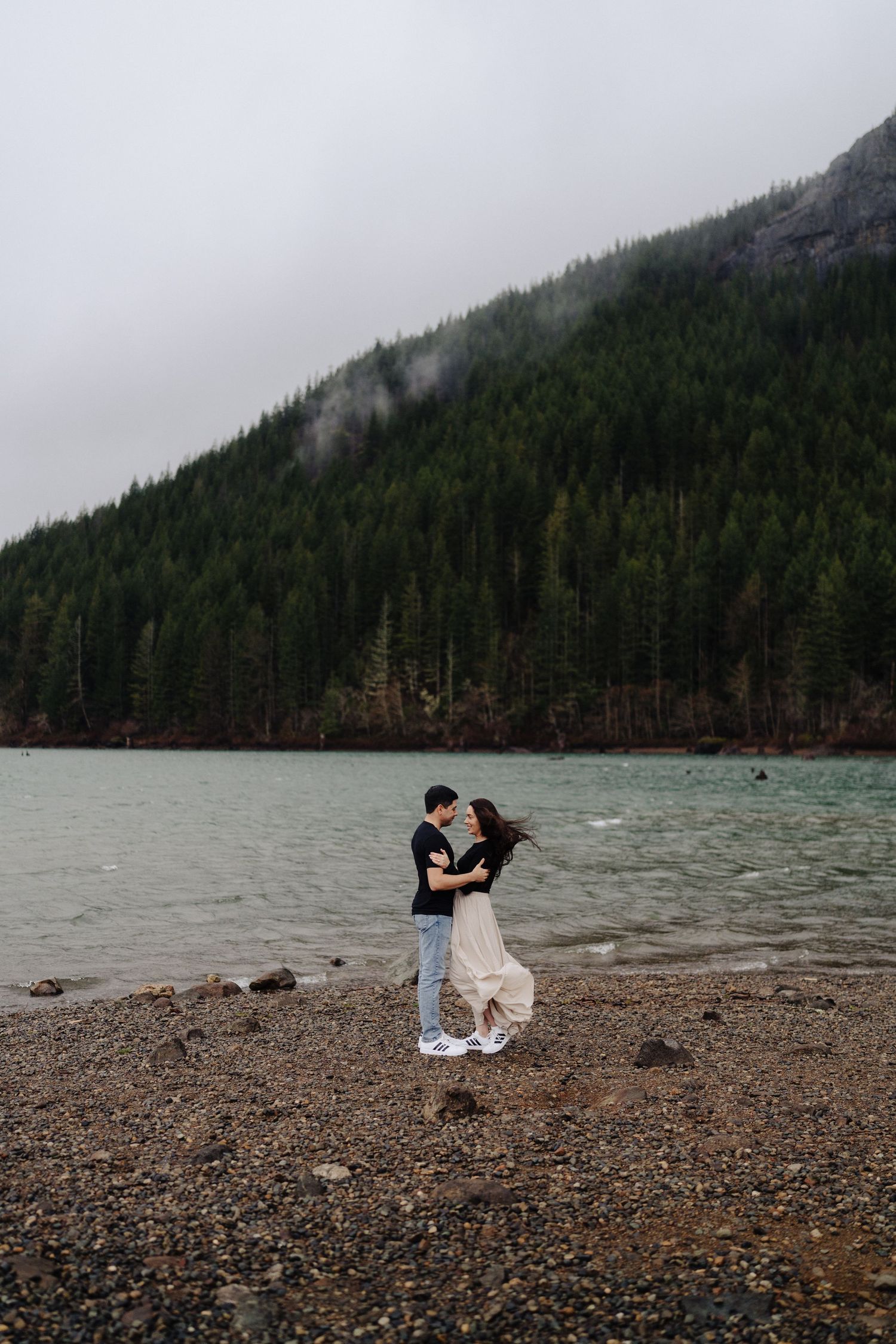 A couple shares an intimate moment on a pebbled shoreline near a misty mountain lake.